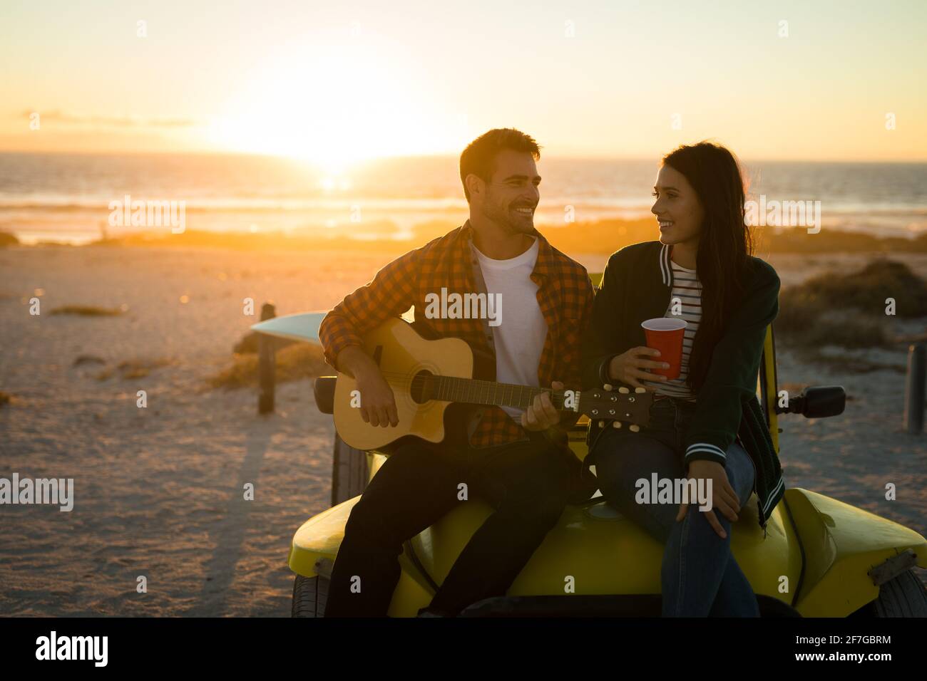 Heureux couple caucasien assis sur la plage buggy au bord de la mer jouer de la guitare et boire au coucher du soleil Banque D'Images