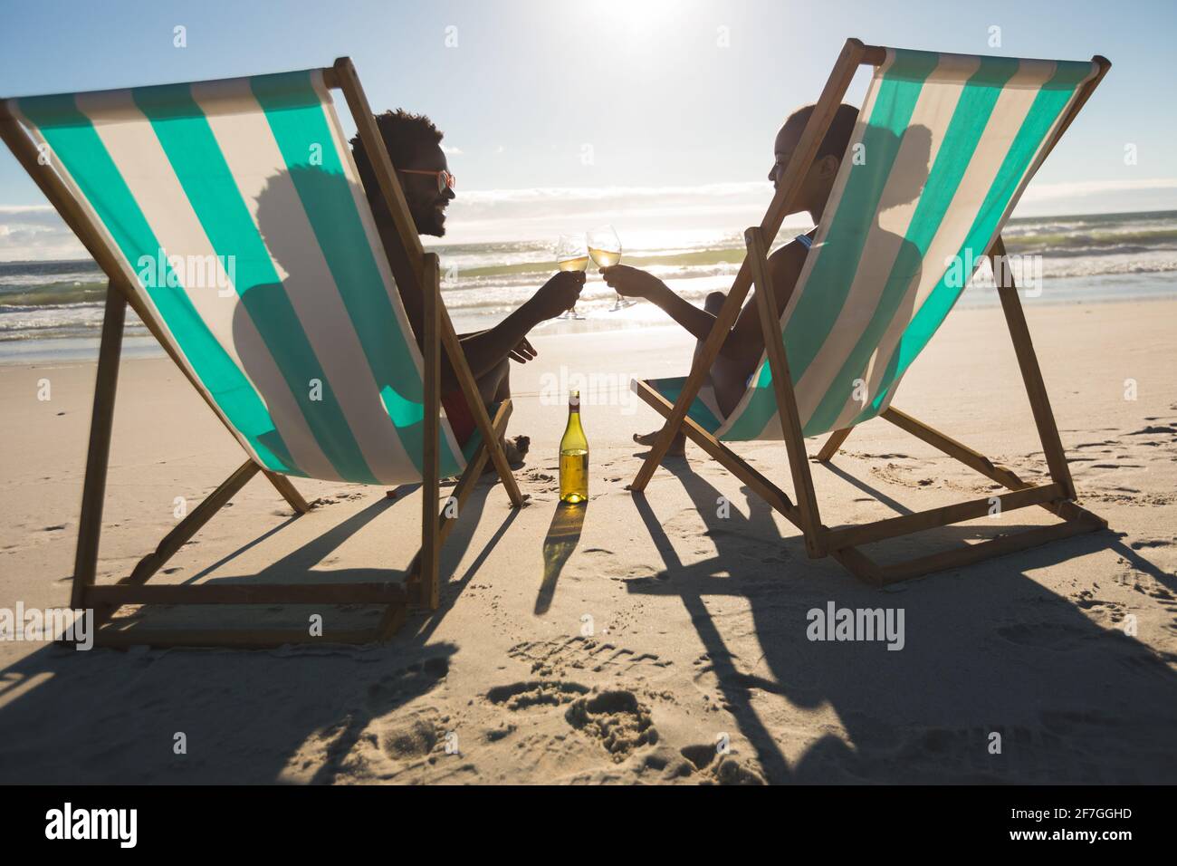 Couple afro-américain amoureux assis dans des chaises longues, prenant des boissons sur la plage Banque D'Images