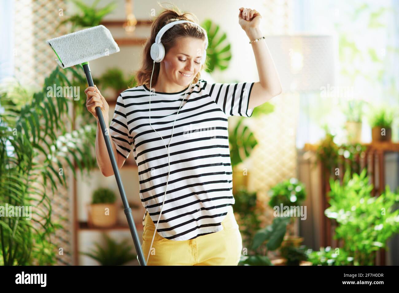 femme d'âge moyen souriante en chemise rayée avec mop et casque faisant des travaux ménagers dans le salon moderne par beau temps. Banque D'Images