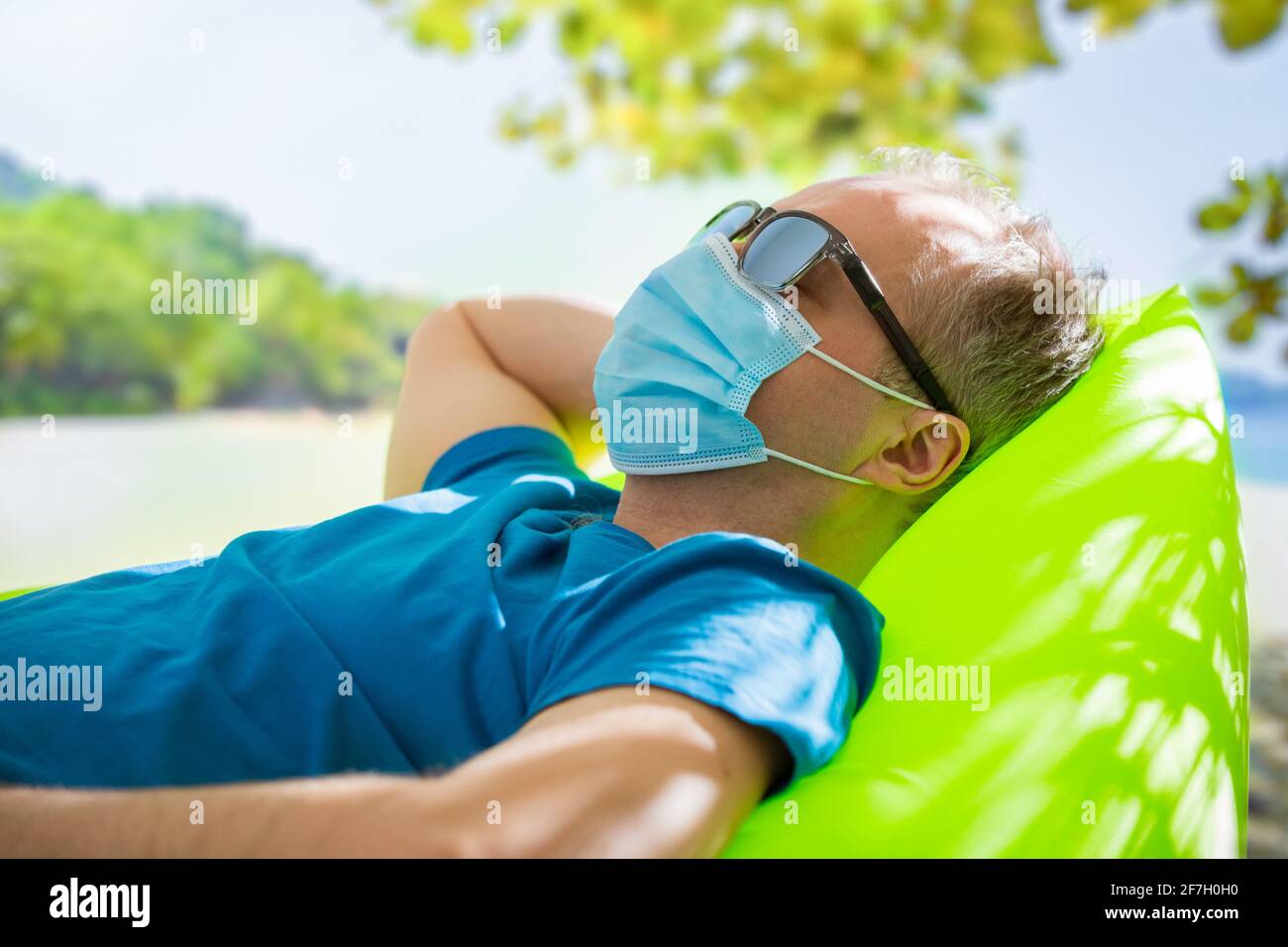 Homme mature dans un masque de protection et des lunettes de soleil se détendant sur la plage. Paysage tropical exotique avec eau turquoise et palmiers. Tourisme en cas de pandémie. Coffre-fort Banque D'Images