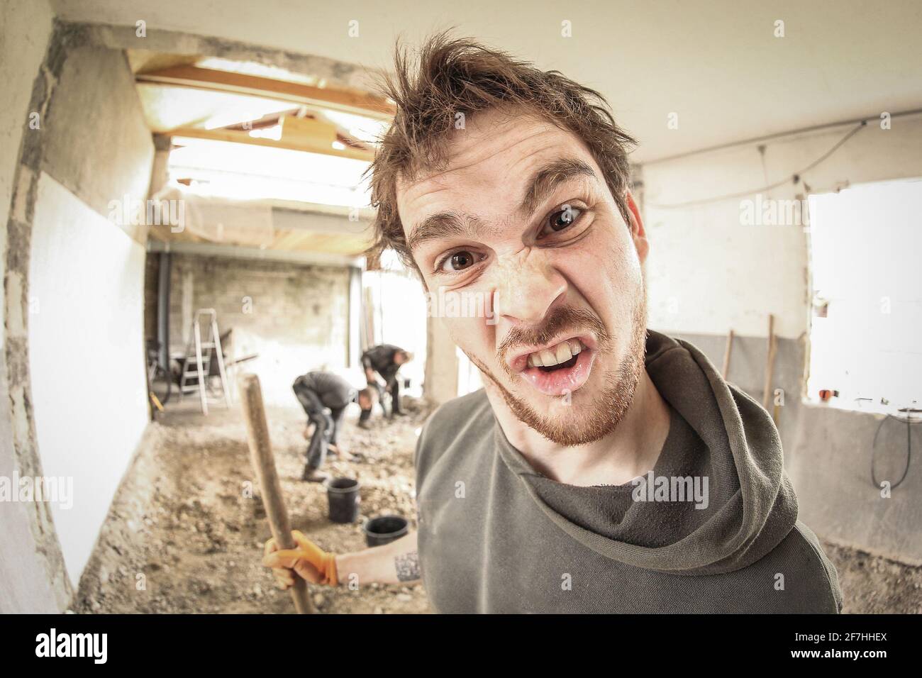 Un travailleur qui fait un visage stupide tout en rénovant une maison. Autres travailleurs en arrière-plan. Vue fisheye. Banque D'Images