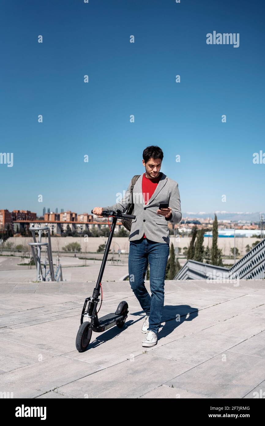 Photo d'un homme d'affaires en veste avec sac à dos se tenir sur un scooter électrique et utiliser un smartphone Banque D'Images