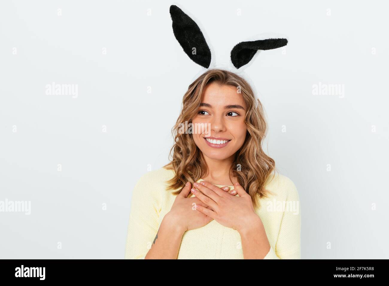 Portrait d'une jeune femme adorable portant des oreilles de lapin de pâques mignonne sourire, en regardant de côté tenant ses mains pliées sur sa poitrine, toile de fond blanche. Banque D'Images