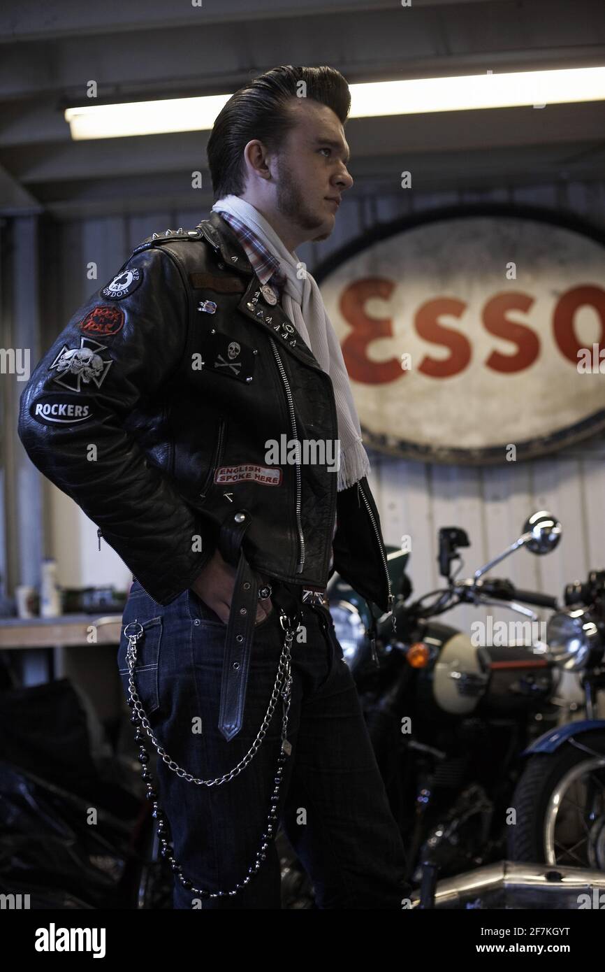 Jeune rocker avec un quiff et une veste en cuir debout dans l'atelier avec le signe Esso en arrière-plan à Londres, Royaume-Uni Banque D'Images
