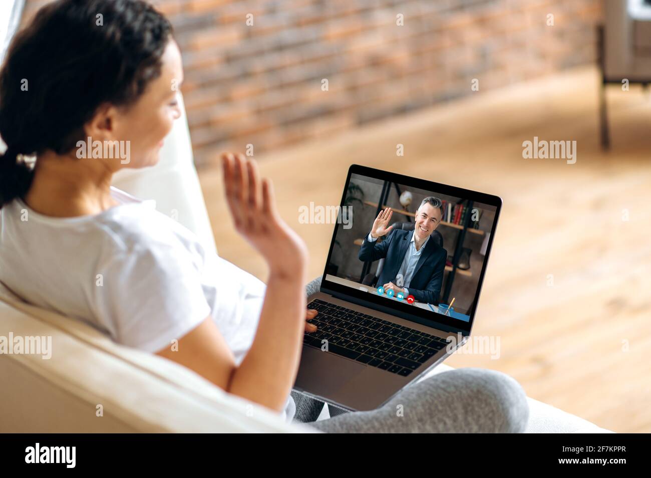 Communication en ligne, visioconférence. Une femme enceinte s'assoit sur un canapé, communique par appel vidéo avec son mari ou collègue bien-aimé à l'aide d'un ordinateur portable, salue l'interlocuteur d'un geste de main Banque D'Images