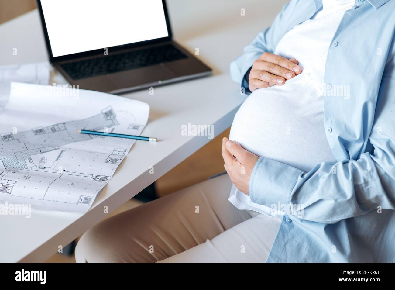 Une femme enceinte, designer ou pigiste, s'assoit à un bureau, épouse doucement son ventre avec ses mains, sur la table un ordinateur portable avec un écran blanc vierge et un document de travail Banque D'Images