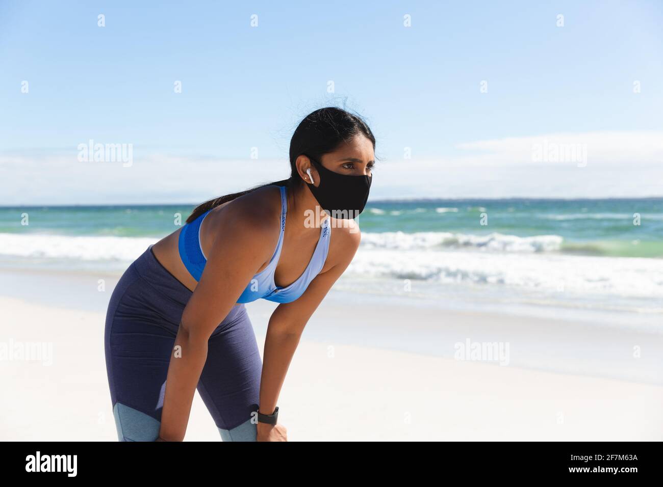 Femme de race mixte s'exerçant sur la plage portant un masque facial et ecouteurs sans fil pour se reposer Banque D'Images