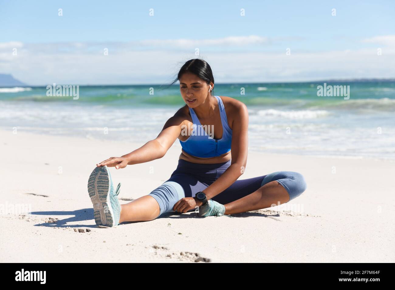 Femme de race mixte s'exerçant sur la plage avec des écouteurs sans fil assis et étirements Banque D'Images
