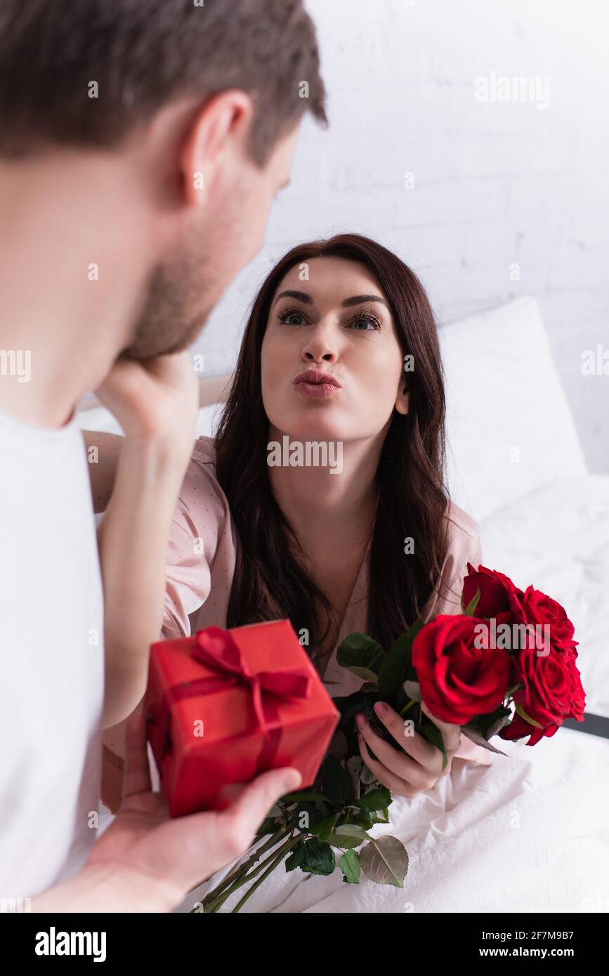 Femme avec fleurs poudant les lèvres près de mari avec le présent sur premier plan flou dans la chambre Banque D'Images