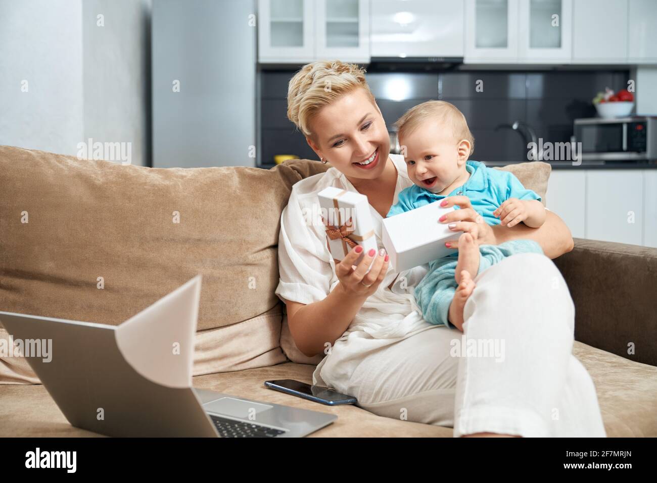 Femme souriante aux cheveux blonds courts jouant avec son petit fils et ouvrant le présent. Ordinateur portable moderne et mobile allongé sur un canapé à proximité. Banque D'Images