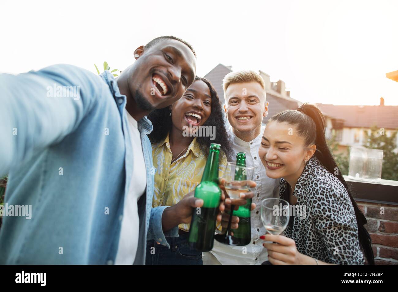 Des amis multiculturels joyeux dans des tenues élégantes emportant le selfie sur un smartphone avec des boissons alcoolisées entre les mains. Concept de personnes, technologie et temps de fête. Banque D'Images