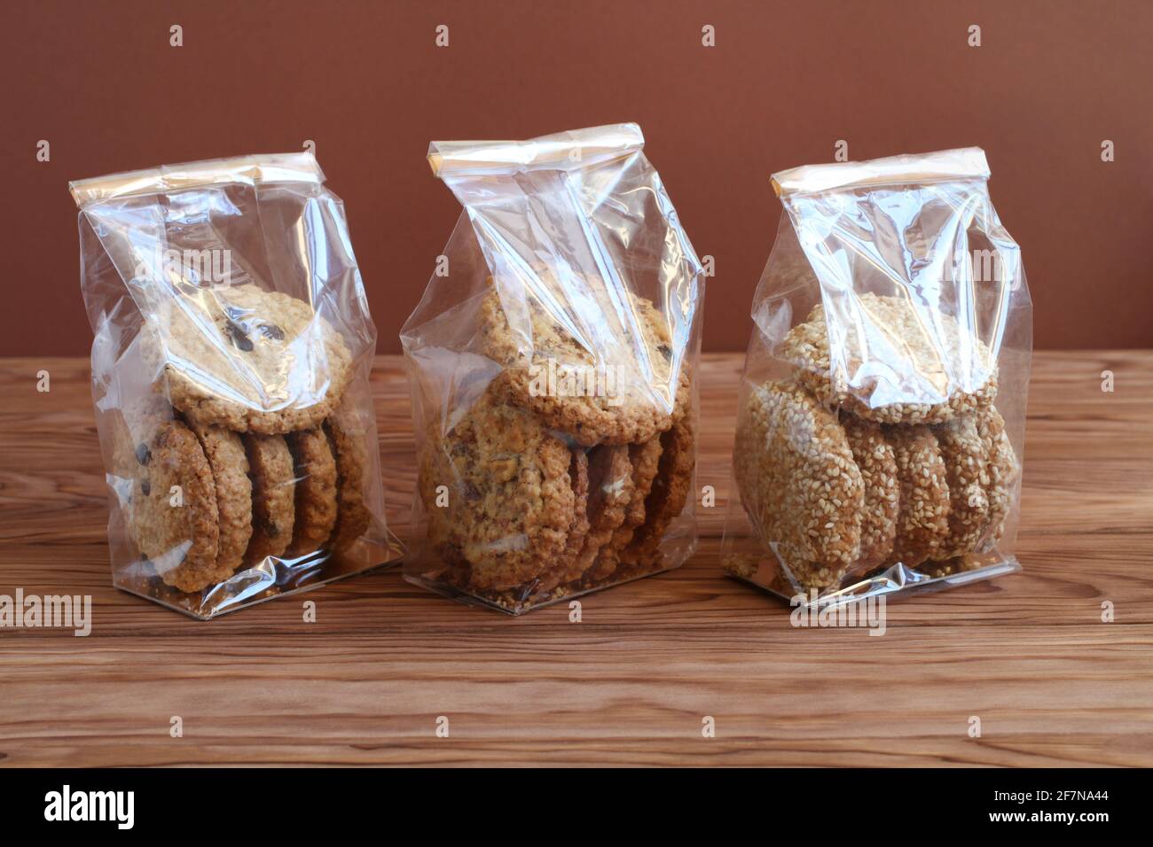 Trois types de biscuits d'avoine - avec chocolat, noix et raisins secs, et graines de sésame - dans des sacs transparents sur une table en bois contre un mur brun. C Banque D'Images