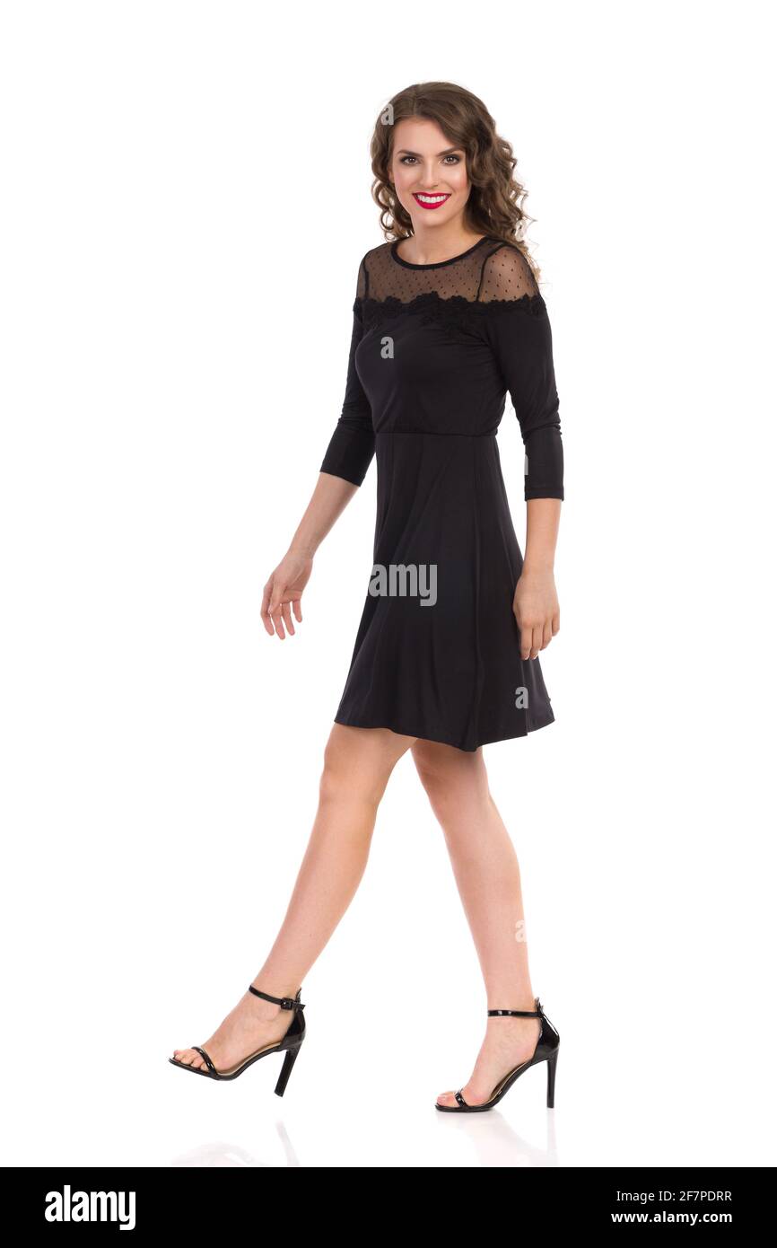 Belle jeune femme en noir élégant robe et talons hauts est de marche et de regarder l'appareil photo et sourire. Vue latérale. Prise de vue en studio pleine longueur isolée Banque D'Images