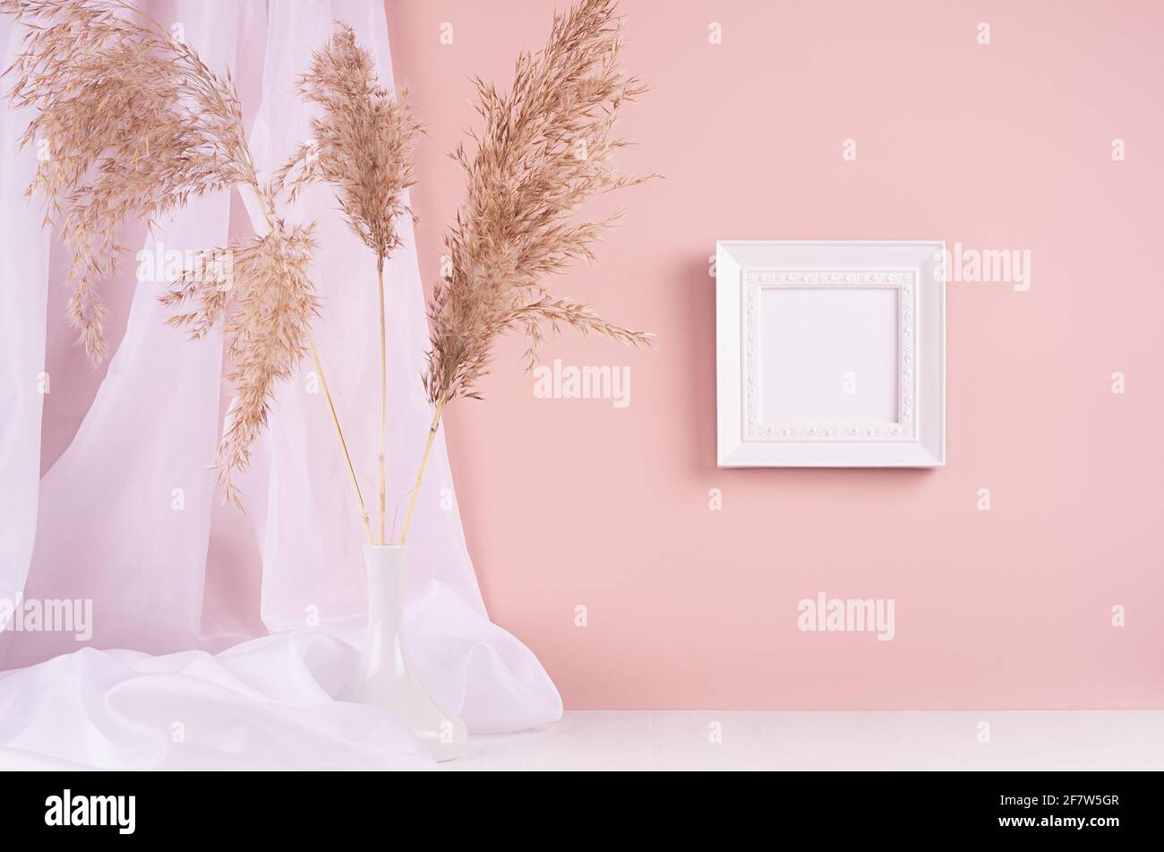 Décoration calme avec cadre photo carré vierge pour le texte suspendu sur un mur rose, rideau en soie, bouquet de roseaux moelleux sur une table en bois blanc. Modèle pour disp Banque D'Images