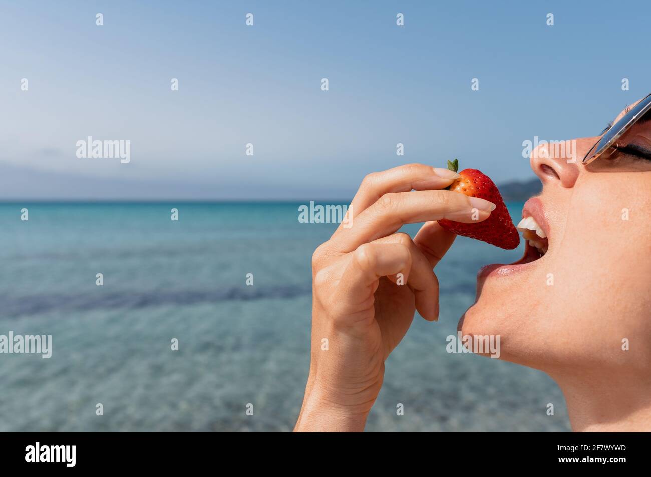 Vue latérale jolie jeune femme mangeant et mordant une fraise mûre avec une  belle mer avec fond d'eau claire Photo Stock - Alamy