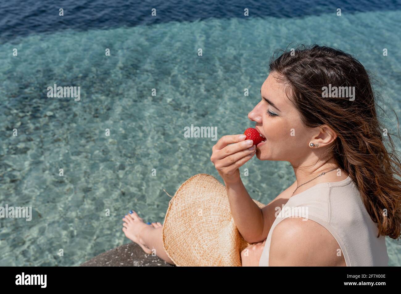 Une jeune femme en gros plan assise sur un rocher mangeant une fraise avec une belle mer transparente translucide sur fond. Le vent qui truffe les cheveux. Banque D'Images