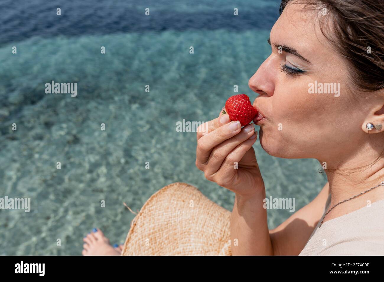 Gros plan jeune femme caucasienne mordant une fraise avec des yeux proches. Magnifique eau de mer translucide sur fond. Banque D'Images