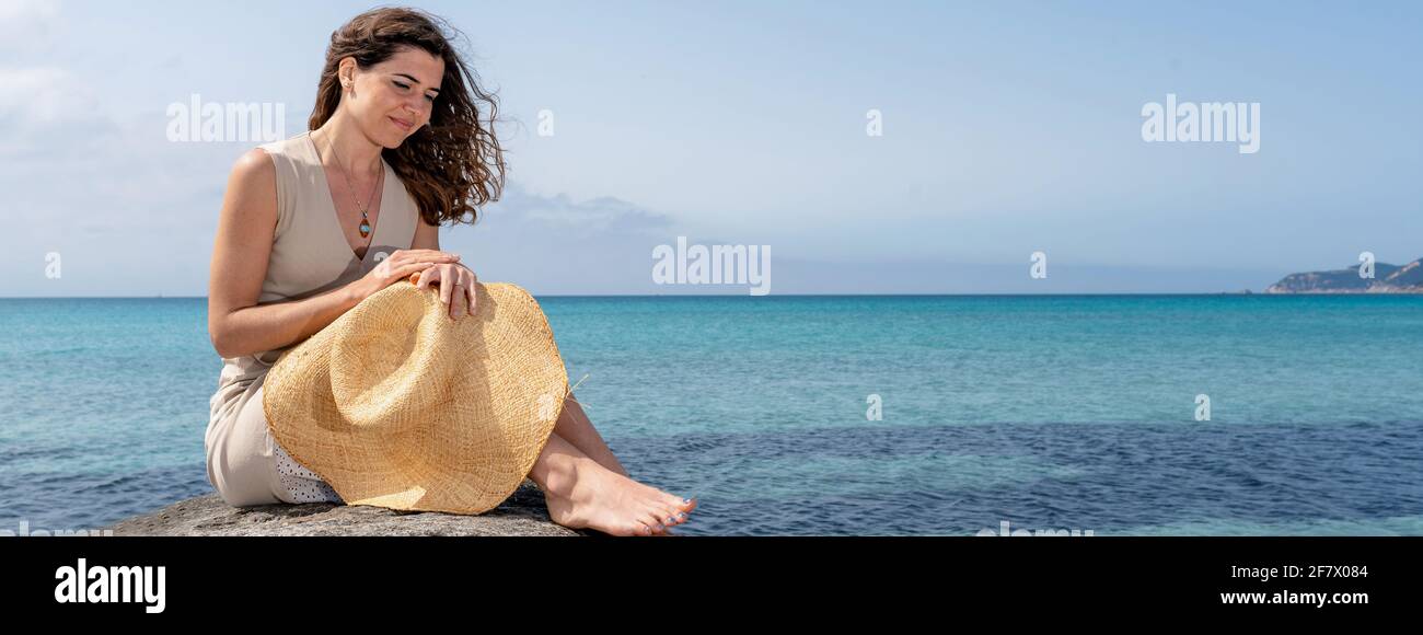 Bannière horizontale ou en-tête avec jeune femme souriante assise sur un rocher avec chapeau de paille au milieu sur la mer. Banque D'Images