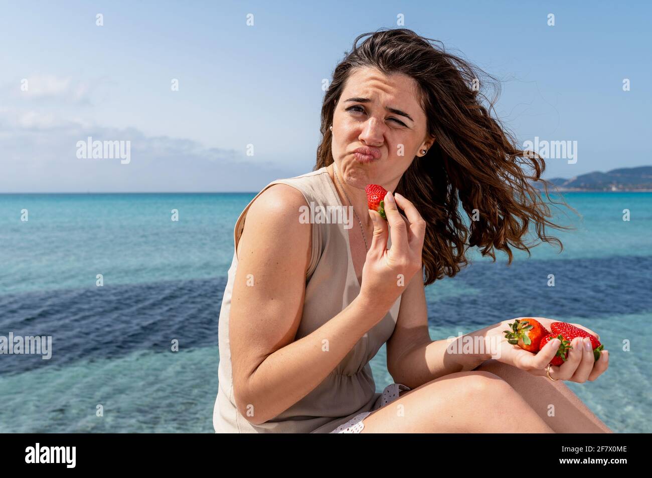 Jeune femme sur la plage avec belle mer sur fond regardant l'appareil photo avec visage drôle tout en mangeant une fraise. Banque D'Images