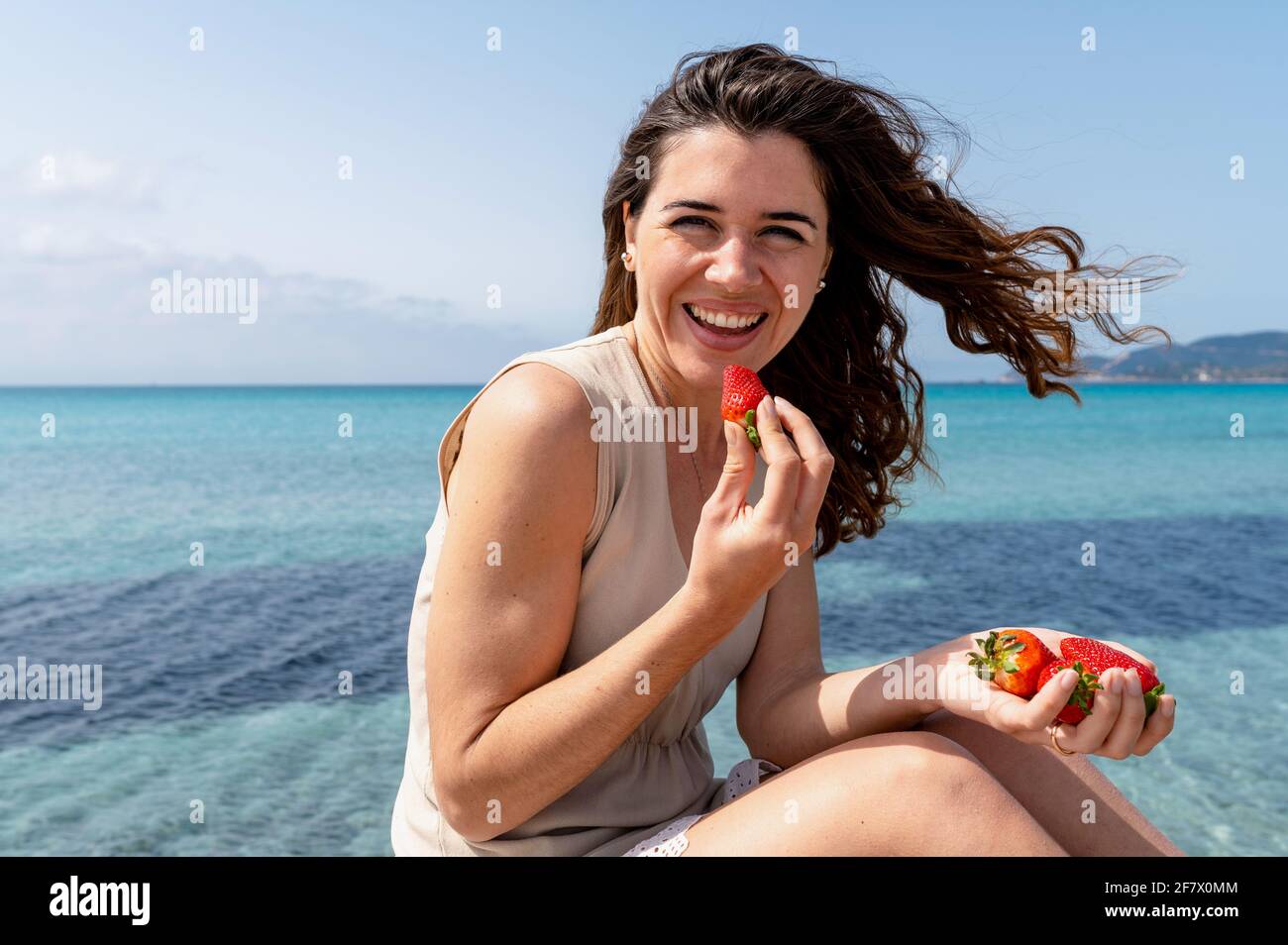 Jeune femme attrayante sur la plage avec belle mer sur fond regardant la caméra avec le visage confiant tout en mangeant une fraise. Banque D'Images