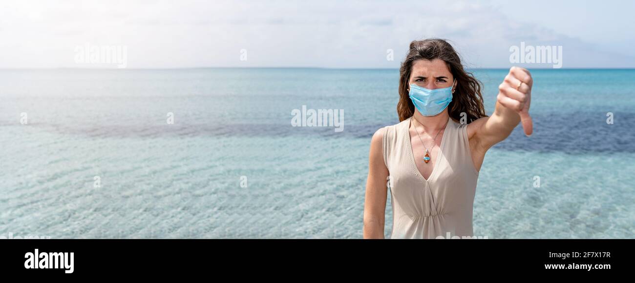 Bannière horizontale ou en-tête Portrait d'une jeune femme portant un masque de protection montrant le pouce vers le bas. Magnifique arrière-plan avec de l'eau bleue claire. Banque D'Images