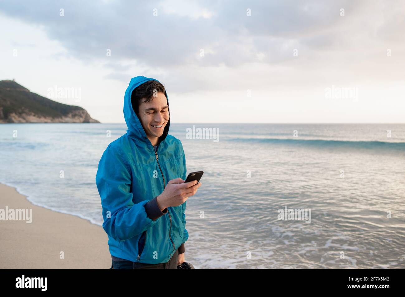 Jeune homme décontracté marchant sur la plage tout en lisant un message texte sur un smartphone et en souriant. Banque D'Images