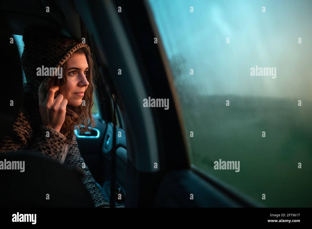 Vue latérale décontractée femme à l'intérieur de la voiture regardant par la fenêtre la pluie tombant avec le coucher de soleil lumière naturelle. Banque D'Images