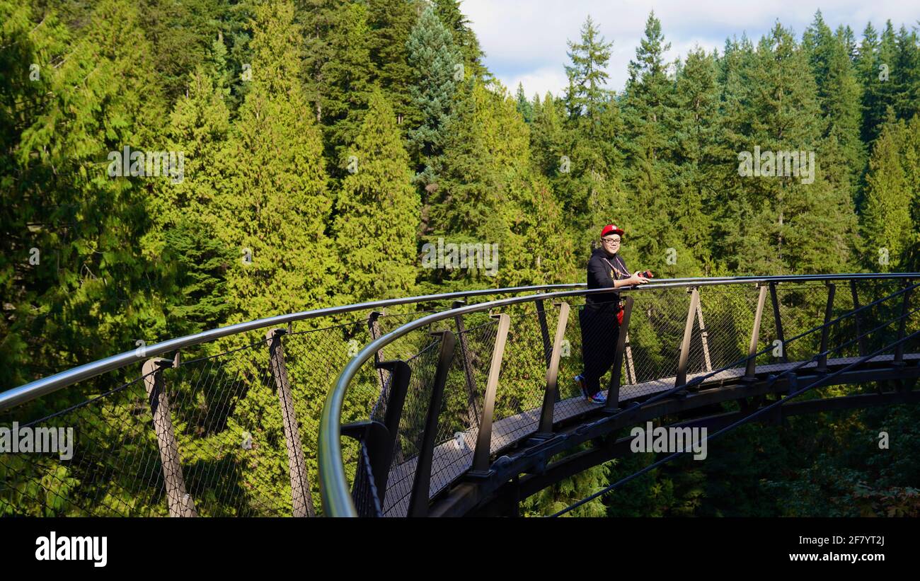 Un jeune homme asiatique en chapeau rouge et en vêtements noirs debout sur un pont aérien, entouré de grands arbres verts dans le parc du pont suspendu Capilano du Canada. Banque D'Images