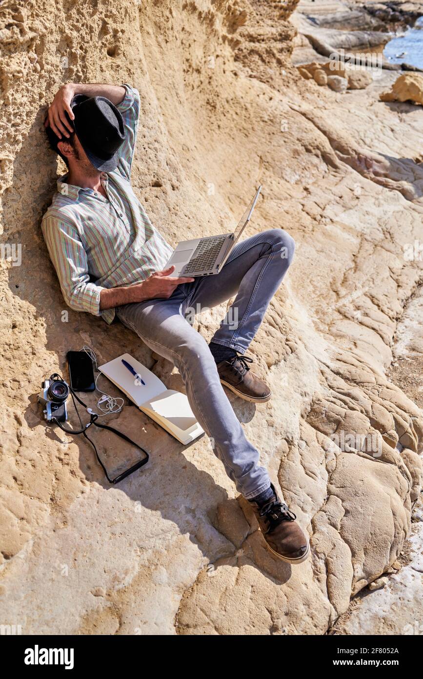 Un jeune homme se détend en mettant son chapeau devant son visage tout en prenant une sieste, il tient son ordinateur portable et à ses côtés il a un appareil photo vintage et un Banque D'Images