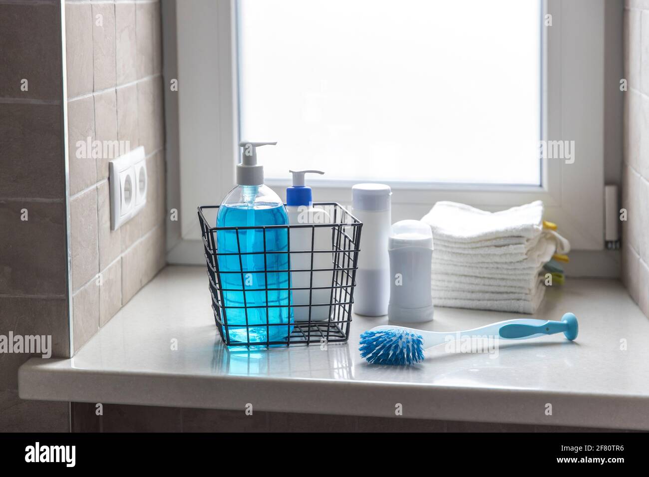 Une bouteille de lotion pour le corps utilisée, du savon liquide bleu se trouve sur un rebord de fenêtre dans une salle de bains avec carreaux beiges. Il y a de petites serviettes de toilette blanches en éponge dans une petite bla Banque D'Images