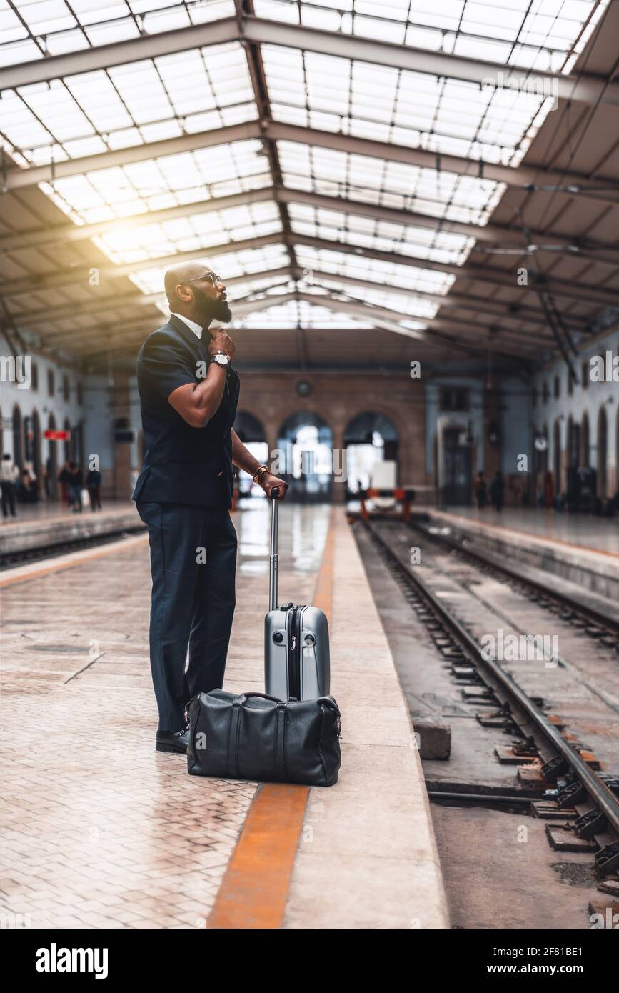 Plan vertical d'un homme d'entrepreneur afro-américain de la dapper ajuster  son cravate en se tenant avec des sacs sur la plate-forme de chemin de fer  de un dépôt de station Photo Stock -