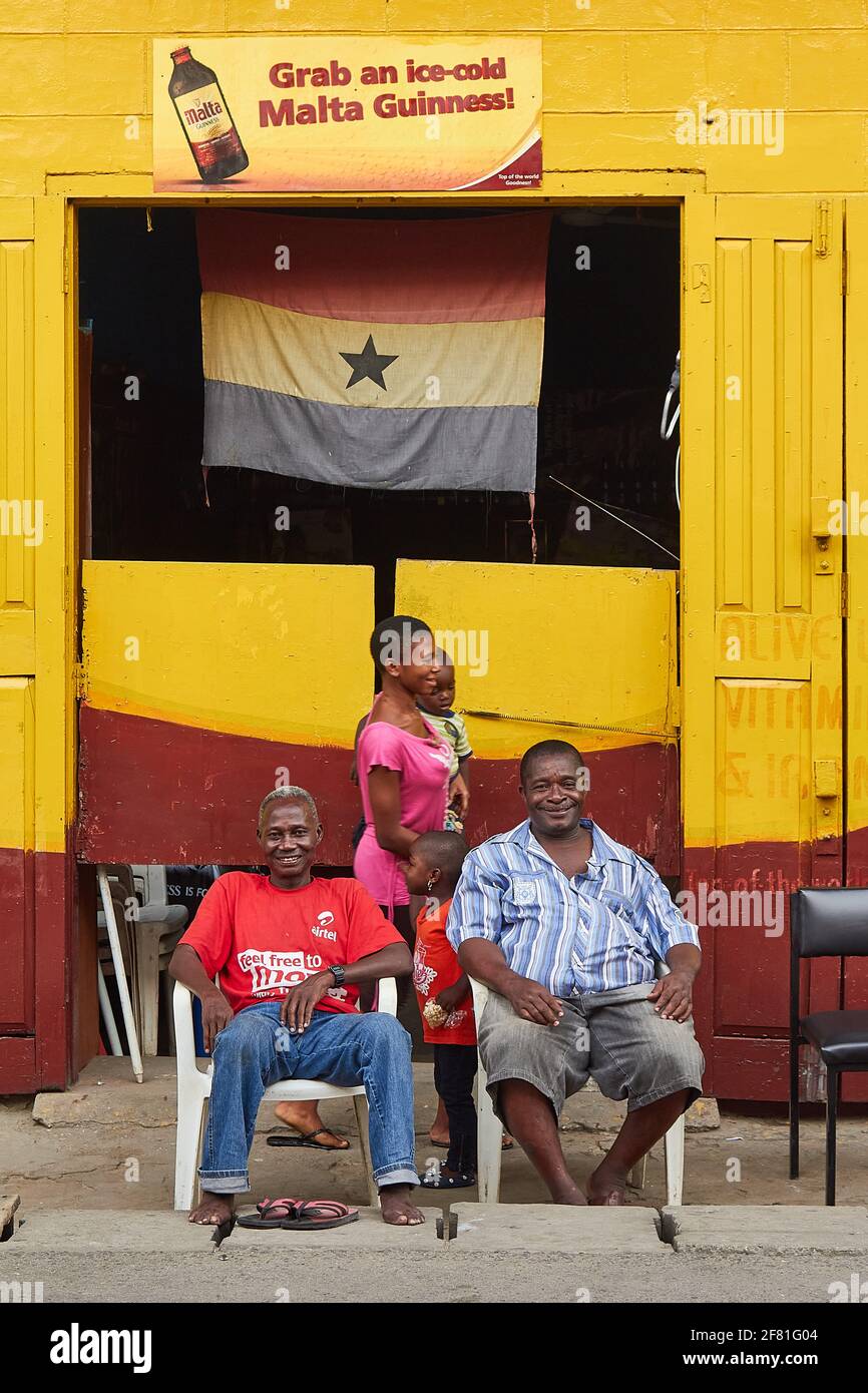 2 hommes assis à l'extérieur d'une barre merveilleusement colorée avec des murs jaunes et un drapeau ghanéen suspendu à la porte de Cape Coast, Ghana Banque D'Images