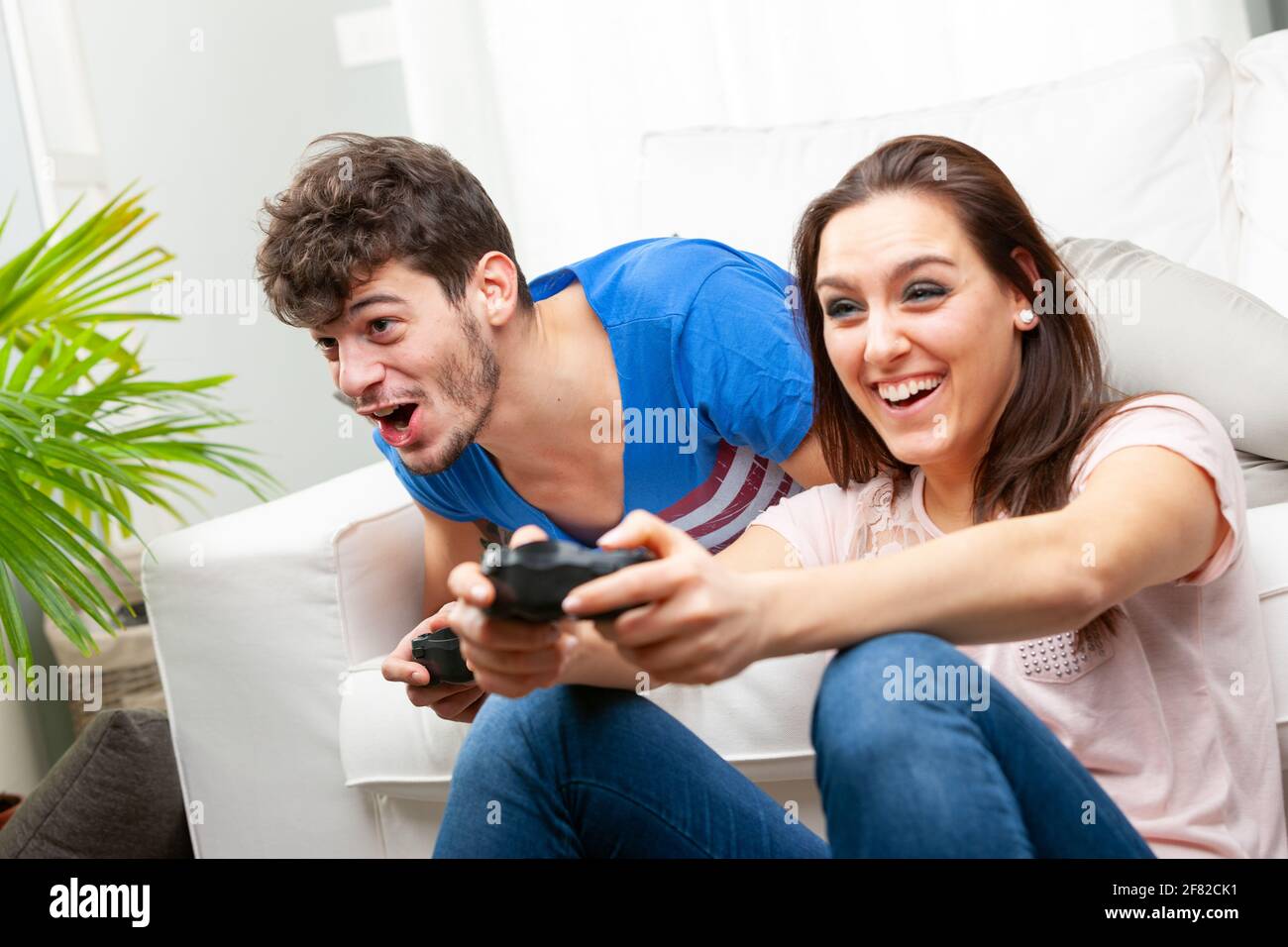 Jeune couple assis sur le plancher tenant les consoles de jeu comme ils jouez ensemble à des jeux vidéo en riant et en hurlant dans leur excitation en gros plan Banque D'Images