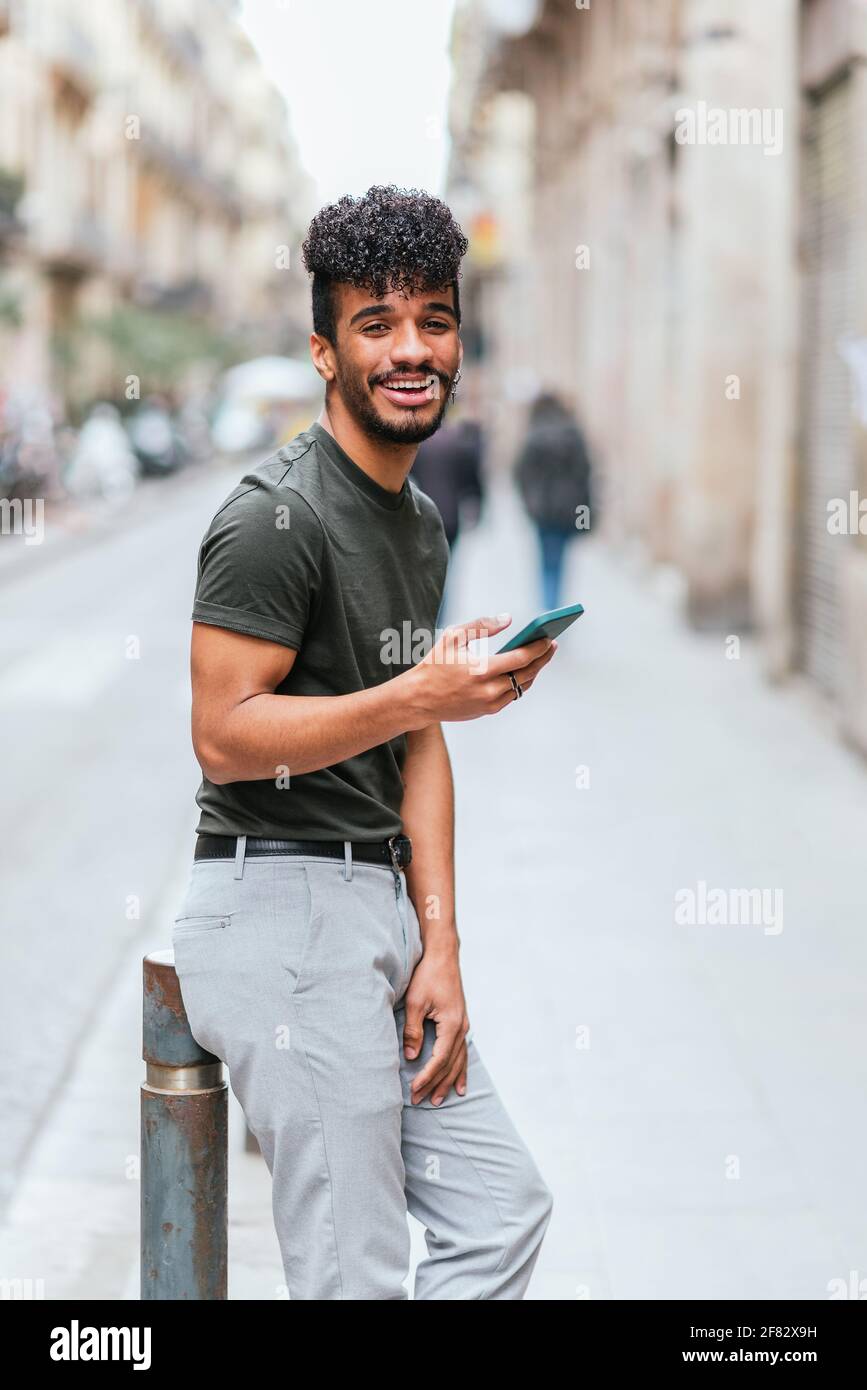 jeune homme hispanique souriant et debout dans la rue regardant la caméra. il porte un t-shirt vert et un jean. Il tient son téléphone. Banque D'Images