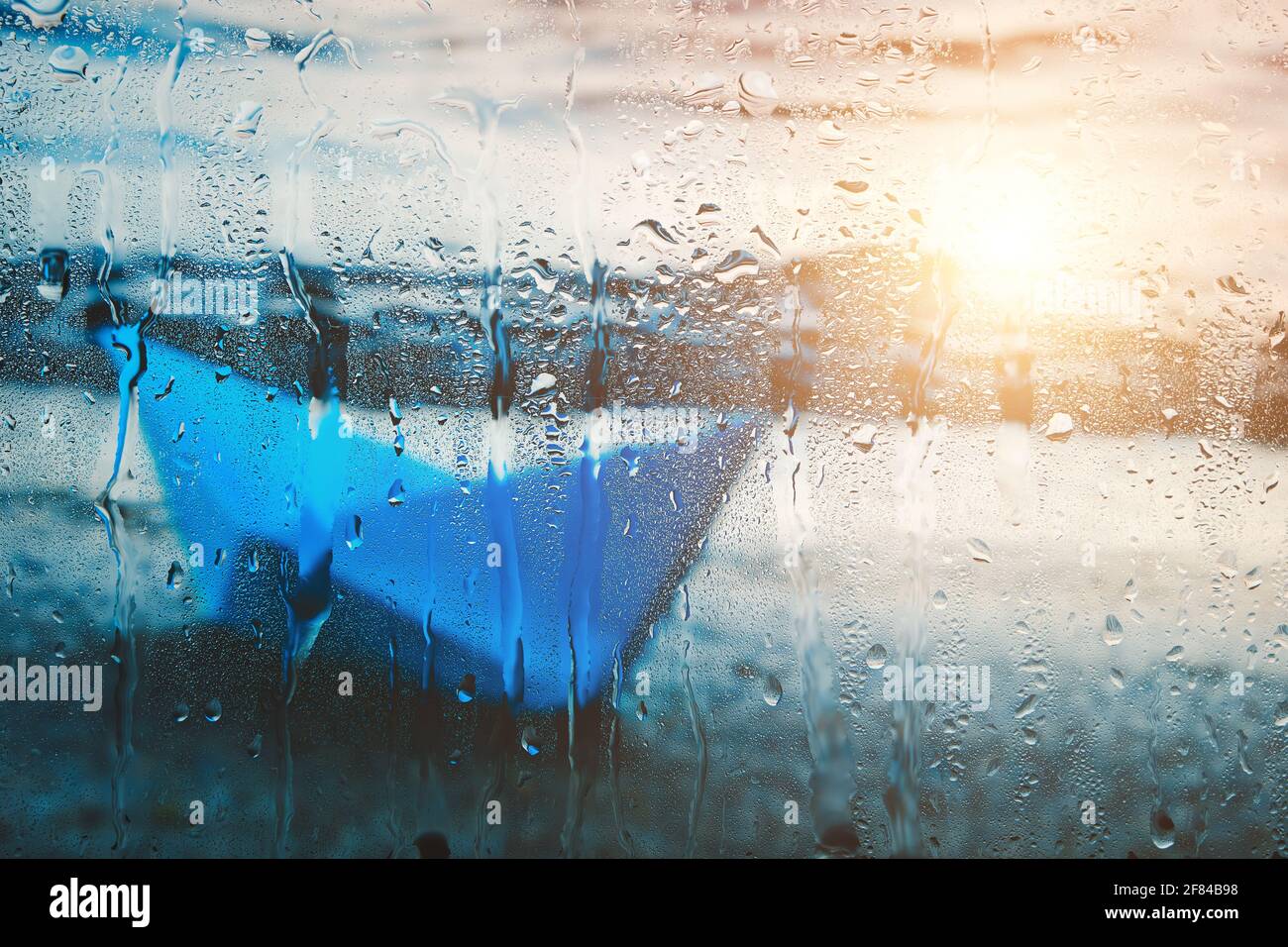 Arrière-plan flou avec lumière du soleil sur l'eau du bateau en papier bleu lavé sur la plage après la pluie derrière le verre humide dedans gouttes de pluie Banque D'Images