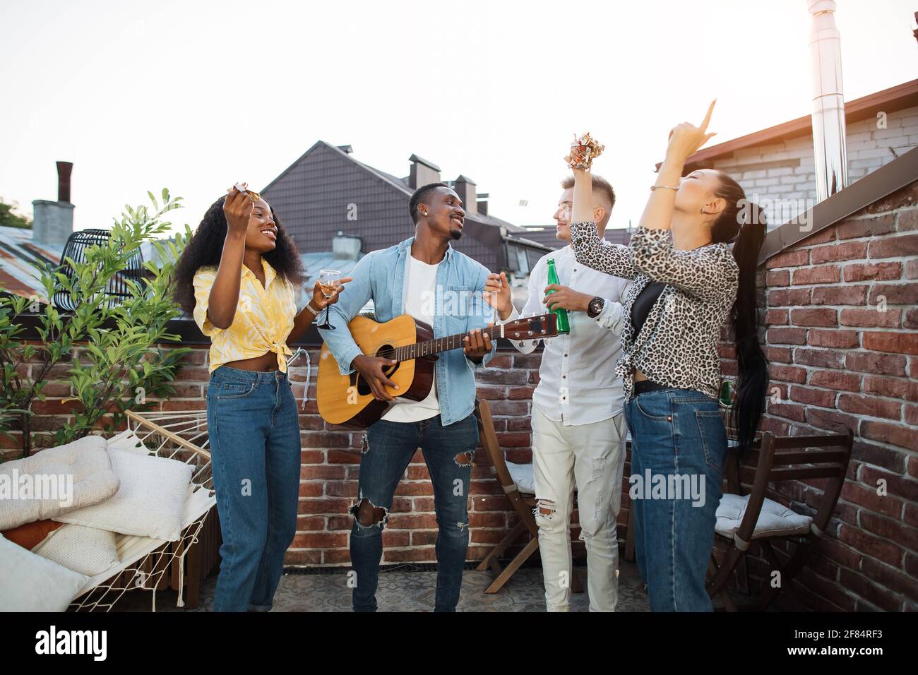 Groupe de quatre personnes multiculturelles en tenues élégantes jouant de la guitare et du chant pendant la fête du toit. Concept d'amitié, de détente et de loisirs. Banque D'Images
