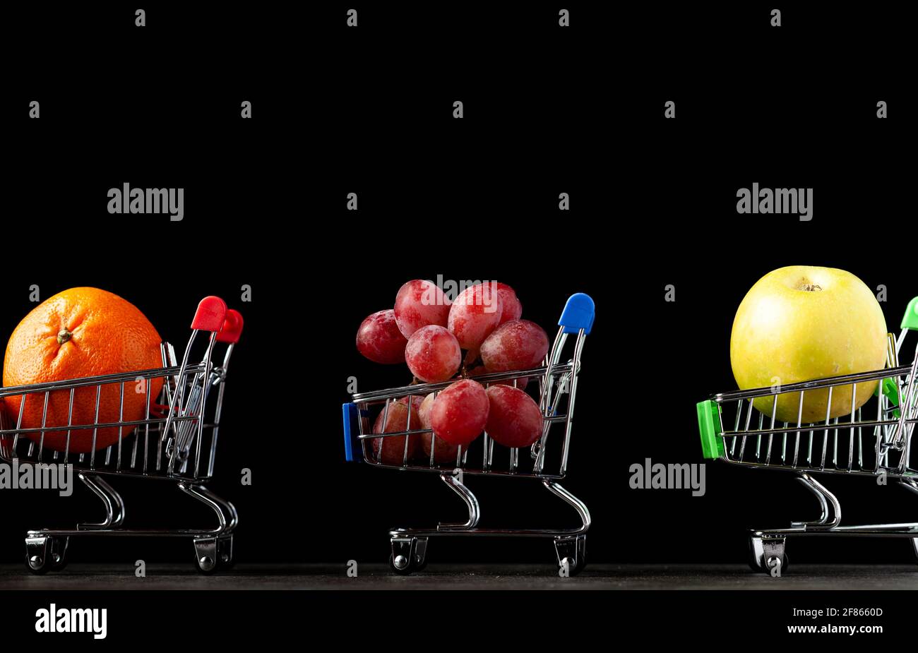 Gros plan, image de fond sombre avec trois chariots de magasinage de jouet rempli de pomme, mandarine et raisins UNE image de concept pour acheter des fruits plus sains a Banque D'Images