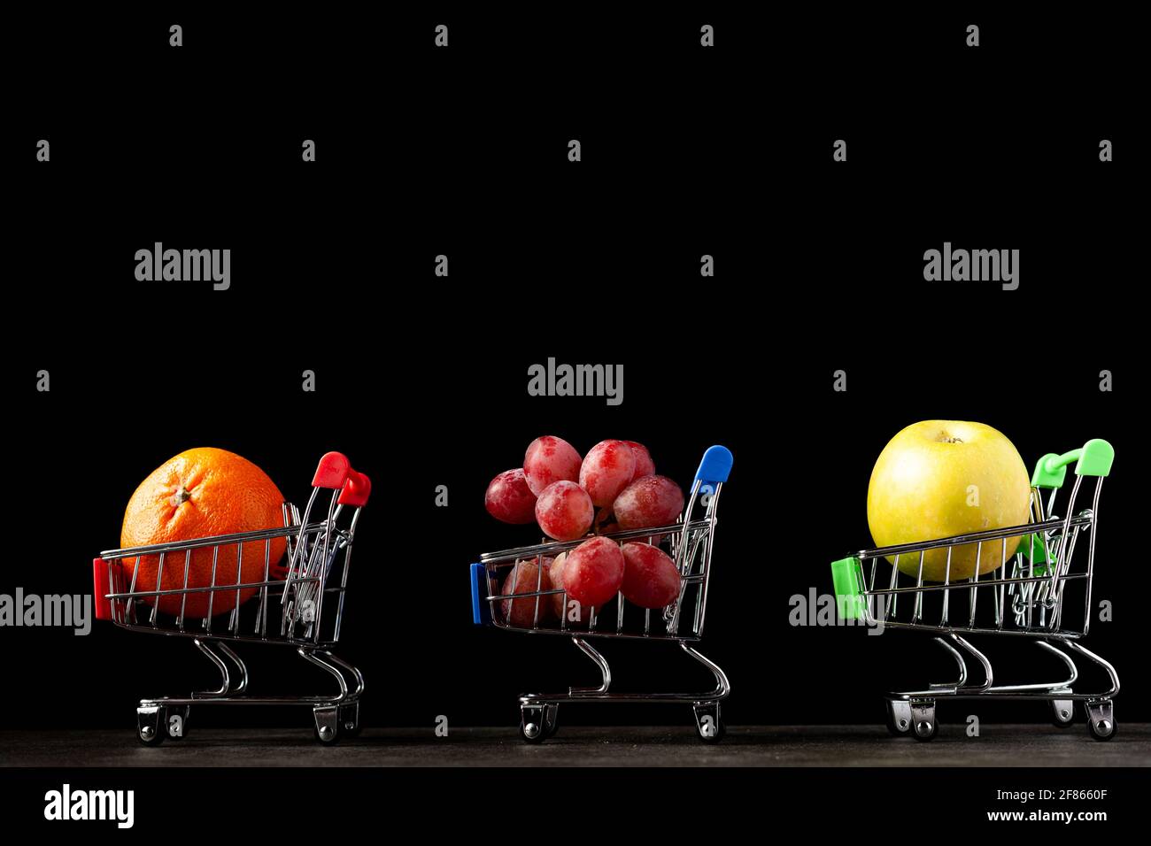 Gros plan, image de fond sombre avec trois chariots de magasinage de jouet rempli de pomme, mandarine et raisins UNE image de concept pour acheter des fruits plus sains a Banque D'Images