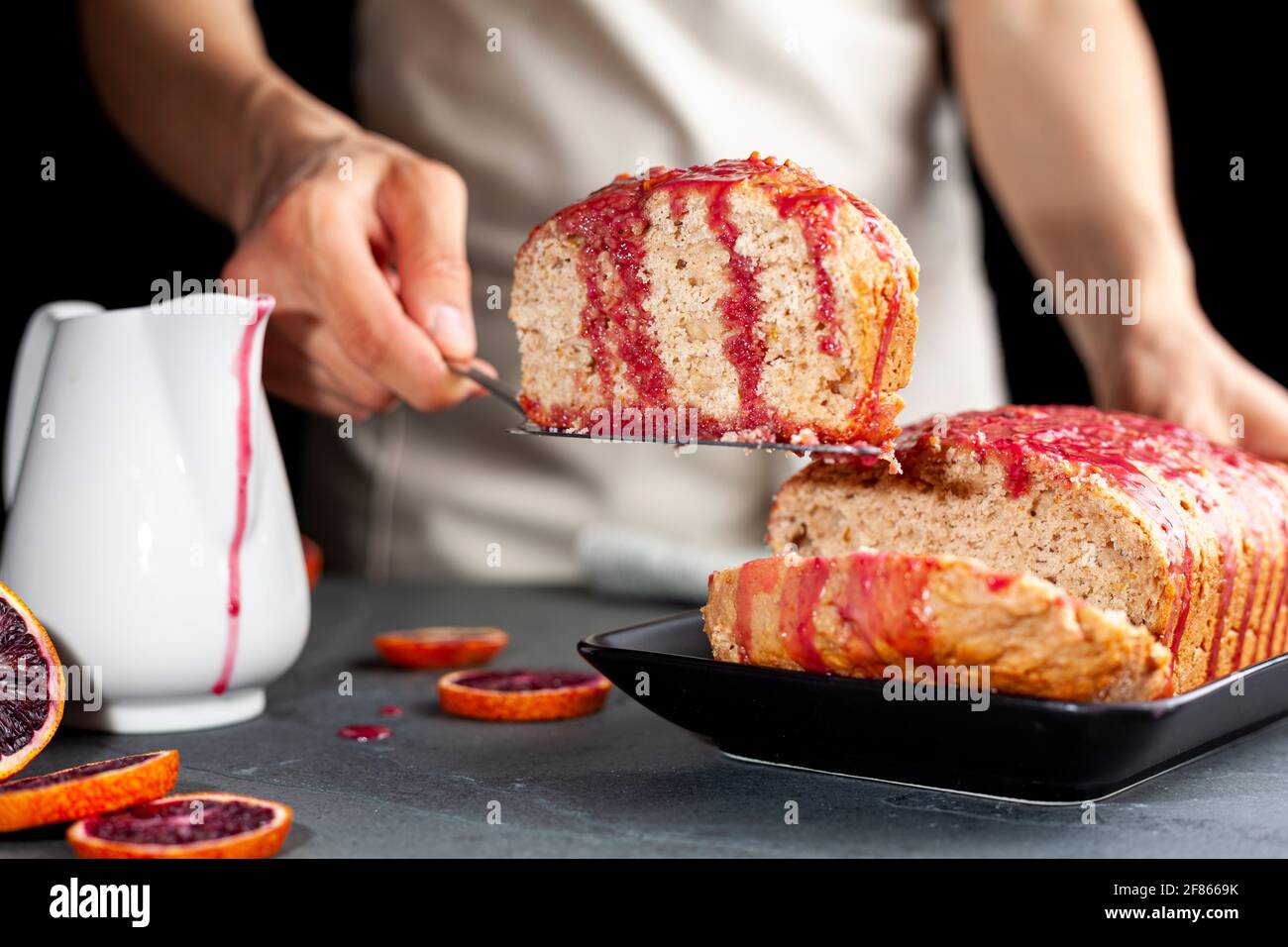 Image en gros plan d'un gâteau d'orange de sang frais fait maison dans un moule à pain et servi sur une assiette en porcelaine noire sur fond de pierre sombre. Une femme sert Banque D'Images