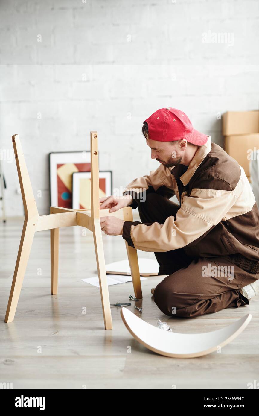 Jeune spécialiste de l'assemblage de meubles debout sur les genoux sur le plancher et l'assemblage d'une nouvelle chaise en bois à l'intérieur du studio, maison ou plat Banque D'Images