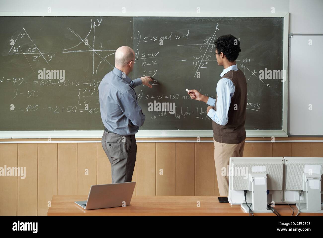 Un professeur bald mature et un jeune étudiant de race mixte discutant de la solution de équation par tableau noir tout en enseignant pointant vers l'un de ses dérivée Banque D'Images
