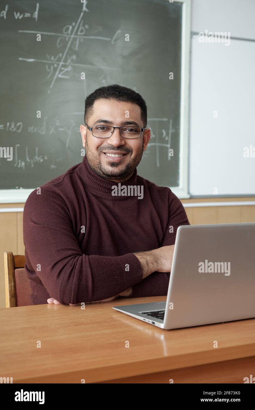 Jeune homme joyeux enseignant de l'université en croisant les bras poitrine assis près du bureau devant l'appareil photo contre tableau noir dans la salle de conférence Banque D'Images