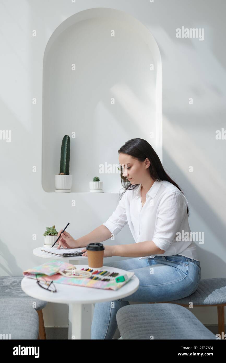 Assez jeune étudiant en art créatif assis à table dans la cafétéria et dessin avec des crayons de couleur dans le carnet de croquis Banque D'Images