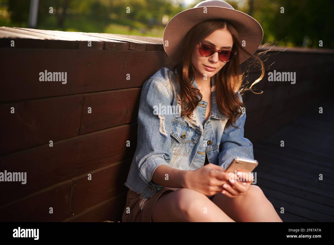 Jolie jeune femme étudiante en lunettes de soleil et veste en denim tendance utilisez votre smartphone en vous asseyant sur une terrasse en bois Banque D'Images