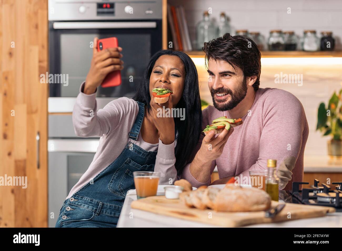 Gaie femme noire et son petit ami ayant délicieux petit déjeuner dans la cuisine à la maison et prenant selfie. Banque D'Images