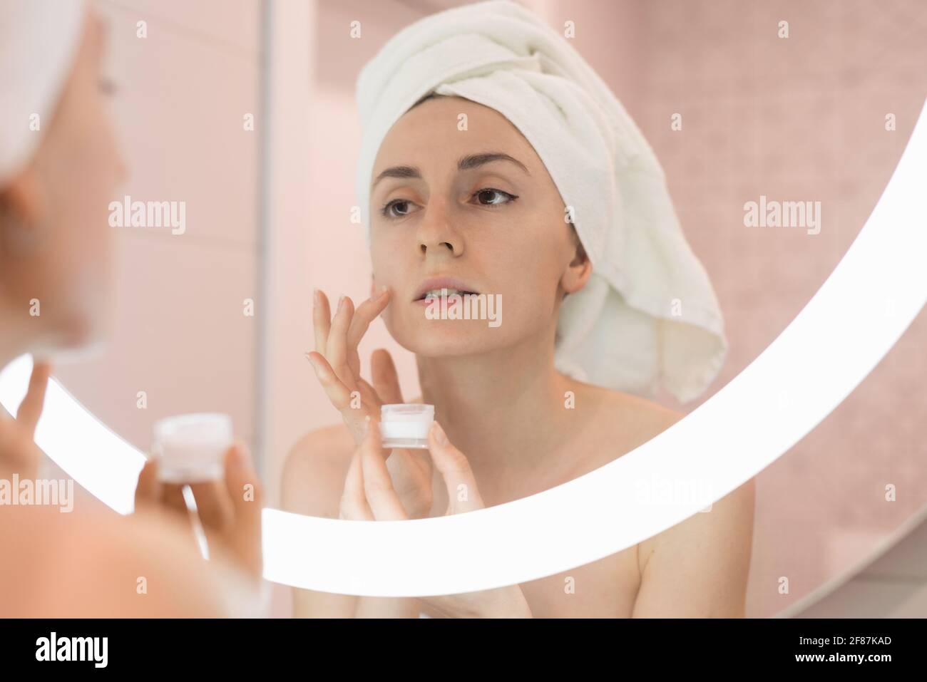 Femme debout près du miroir dans la salle de bains et appliquant un hydratant sur son visage Banque D'Images