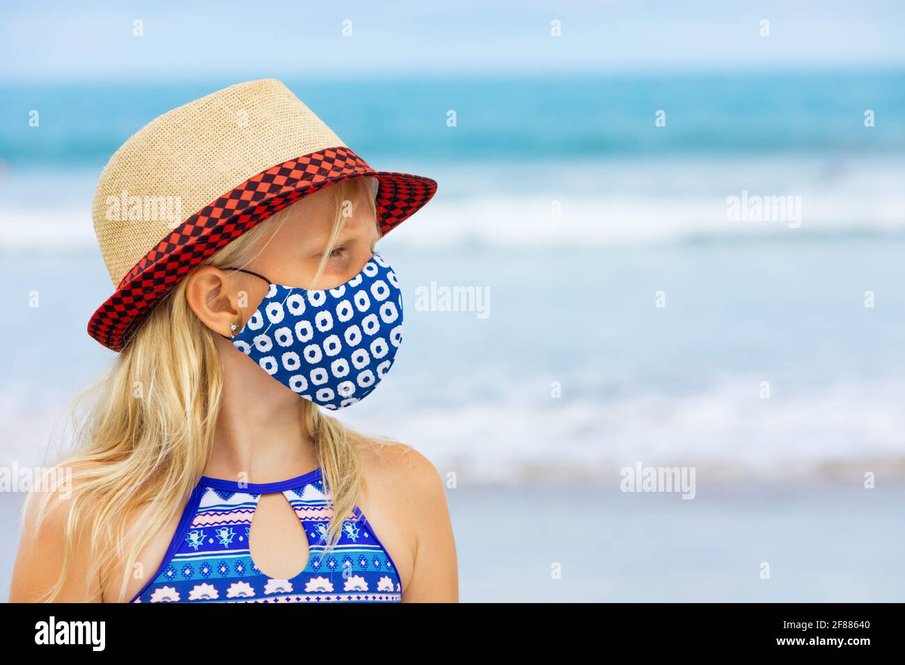 Enfant en chapeau de paille, les masques élégants s'amusent sur la plage. Nouvelles règles pour le port de couvre-visage dans les lieux publics. Nouvelles règles pour porter un masque de couverture en tissu Banque D'Images