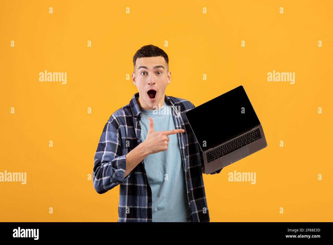 Un jeune homme choqué pointe vers un écran d'ordinateur portable vide avec de l'espace pour la conception de sites web sur fond orange studio Banque D'Images