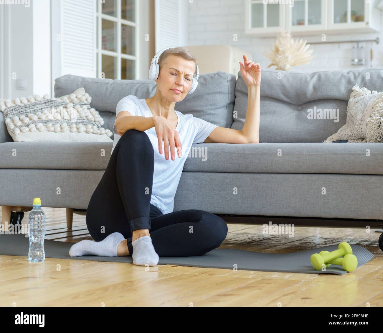 Femme âgée avec des yeux fermés dans un casque d'écoute écouter de la musique dans le salon. Détendez-vous après avoir fait de l'exercice. Santé émotionnelle et spirituelle. Bien-être, bien-être avec méditation. Profitez de la vie lente Banque D'Images