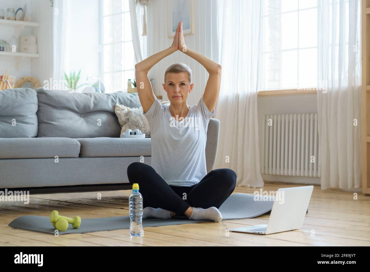 Femme âgée aux yeux fermés, formation en ligne dans le salon. Faites de l'exercice avec un ordinateur portable et suivez des cours d'entraînement. Santé émotionnelle et spirituelle. Bien-être, bien-être avec méditation. Profitez de moments inoubliables Banque D'Images